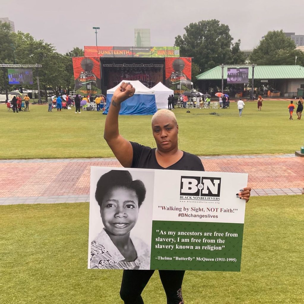Mandisa Thomas, the founder of Black Nonbelievers, stands with her right fist raised in the air and the left hand holding a sign. The sign has the Black Nonbelievers logo, their slogan "walking by sight, not faith", a picture of Thelma "Butterfly" McQueen, and a quote from her saying "As my ancestors are free from slavery, I am free from the slavery known as religion."